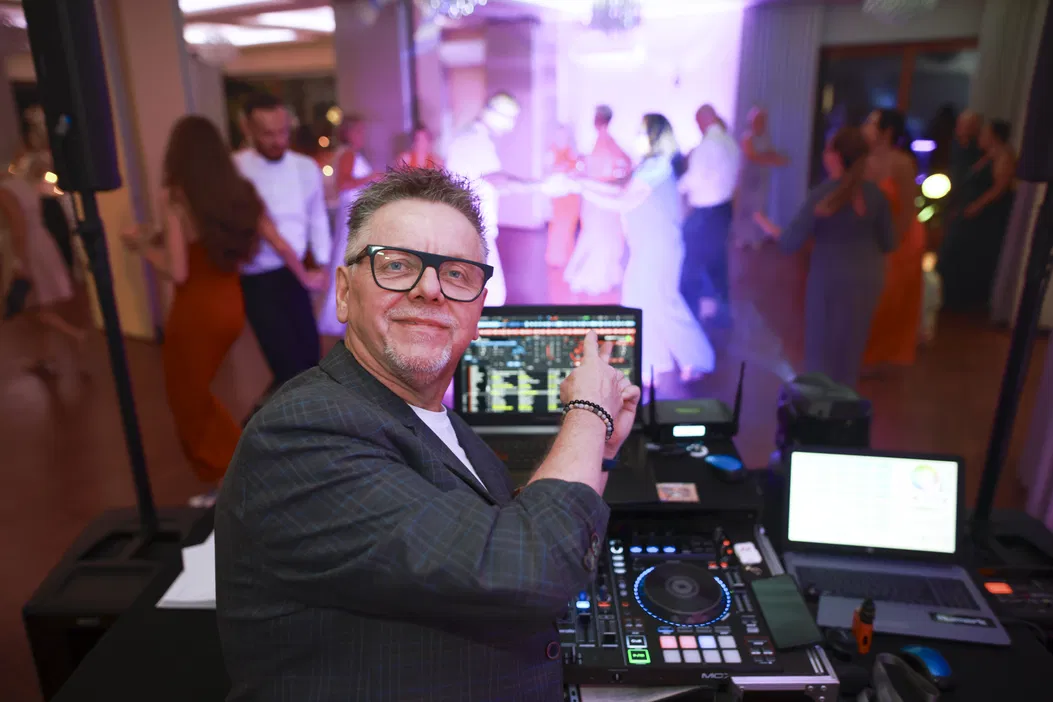 DJ na weselu, ludzie tańczący w tle, sprzęt DJ-ski, muzyka na żywo, przyjęcie, impreza taneczna.
