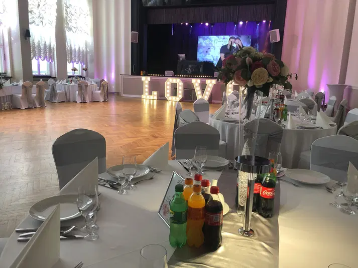 Sala weselna z elegancko nakrytymi stołami, dekoracjami kwiatowymi i dużym napisem LOVE na scenie. 