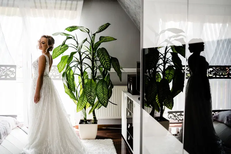  Panna młoda w białej sukni ślubnej stojąca obok dużej rośliny w jasnym pokoju z eleganckim wystrojem. 