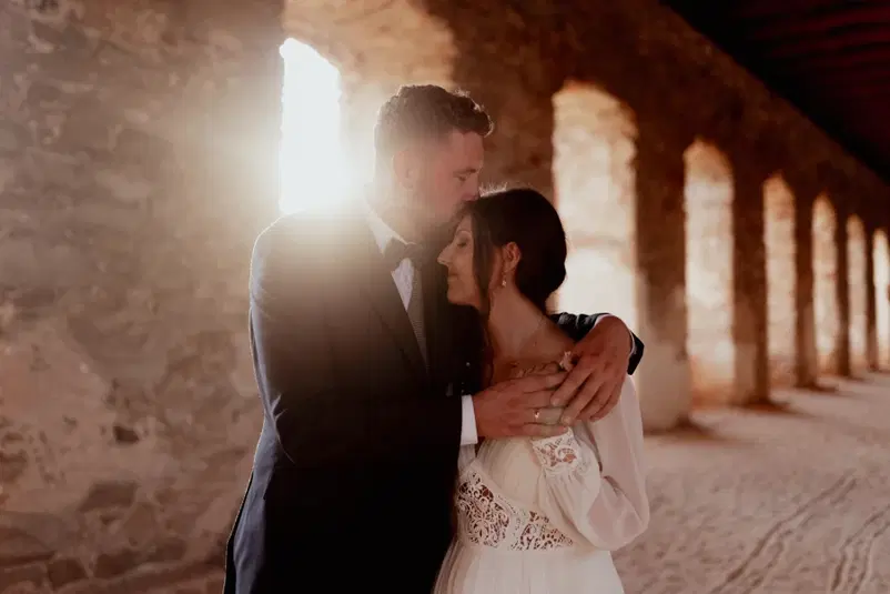  Para w eleganckich strojach ślubnych w romantycznym uścisku w zabytkowym budynku, światło zachodzącego słońca tworzy magiczną atmosferę. 