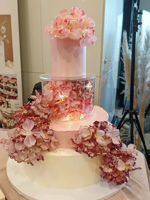 Tort weselny z trzema poziomami ozdobiony różowymi kwiatami hortensji i delikatnymi światełkami LED, elegancki design, doskonały na luksusowe przyjęcia.