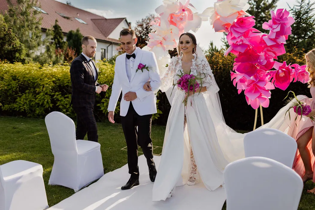 Ślub na świeżym powietrzu, para młoda na ceremonii, dekoracje z dużych kwiatów, eleganckie stroje, bukiet z różowymi kwiatami.