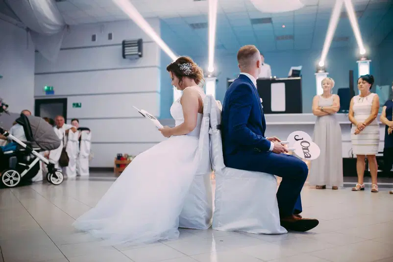  Para młoda podczas zabawy weselnej, siedząca tyłem do siebie na krzesłach w sali weselnej, w otoczeniu gości. 