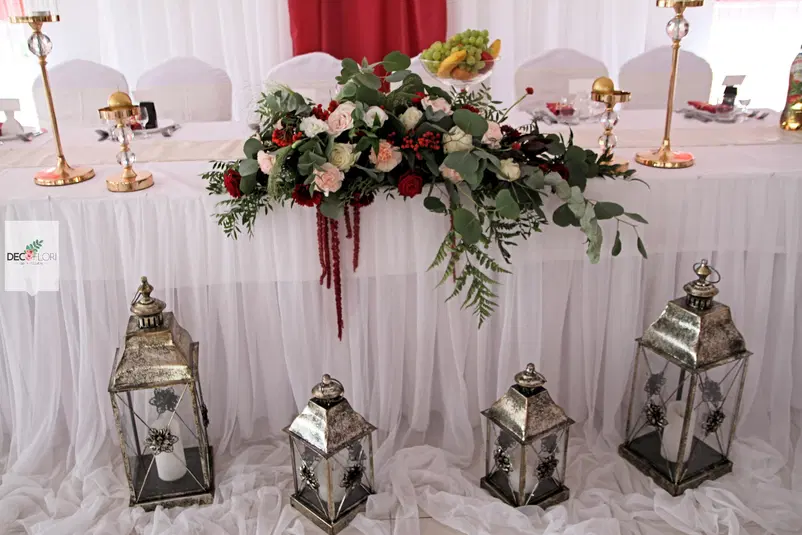  Elegancka dekoracja stołu weselnego z kwiatowym bukietem i latarniami, złote świeczniki, biała tkanina, wesele, dekoracje stołu, aranżacja kwiatowa. 