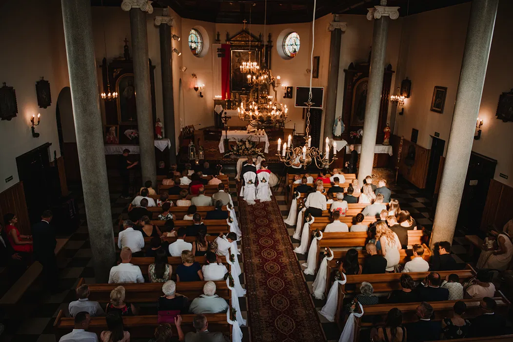 Wnętrze kościoła podczas ceremonii ślubnej, widok z góry na gości siedzących w ławkach, ołtarz ozdobiony kwiatami, żyrandole zwisające z sufitu.