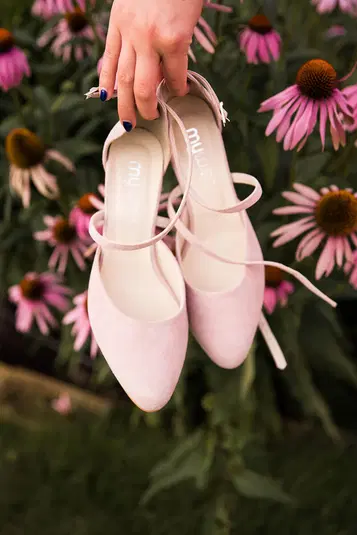  Różowe eleganckie buty damskie z paskami trzymane w dłoni na tle kwiatów jeżówki. 