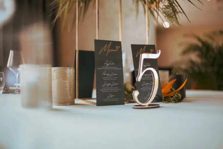  Stylowa dekoracja stołu weselnego z eleganckim menu i numerem stolika otoczonym roślinami. 