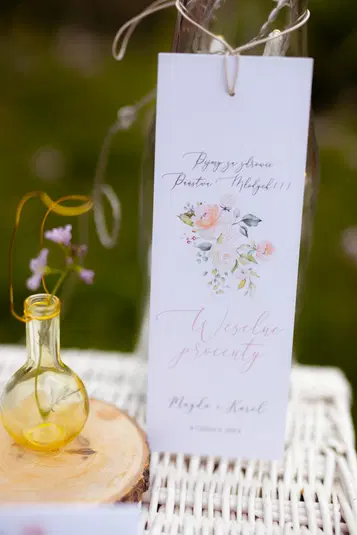  Karta z podziękowaniem weselnym z napisem Weselne procenty ozdobiona florystycznym motywem, stojąca na wiklinowym tle obok ozdobnej żółtej butelki. 