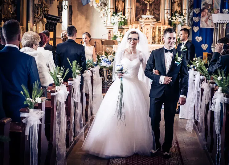  Ślub w kościele, para młoda wychodzi z ceremonii, eleganckie stroje, dekoracje kwiatowe, panna młoda w białej sukni, pan młody w czarnym garniturze, goście zgromadzeni w tle. 