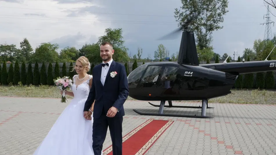  Para młoda w eleganckich strojach ślubnych stoi na tle czarnego helikoptera, trzymając się za ręce i uśmiechając, z bukietem kwiatów i czerwonym dywanem na podjeździe. 
