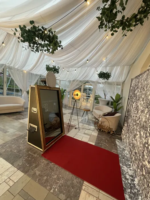 Elegancki wystrój wnętrza z lustrem fotograficznym na czerwonym dywanie, białe draperie pod sufitem, lampki i rośliny tworzące romantyczną atmosferę. Idealne miejsce na wesela i przyjęcia, nowoczesna aranżacja, fotolustro na imprezy.