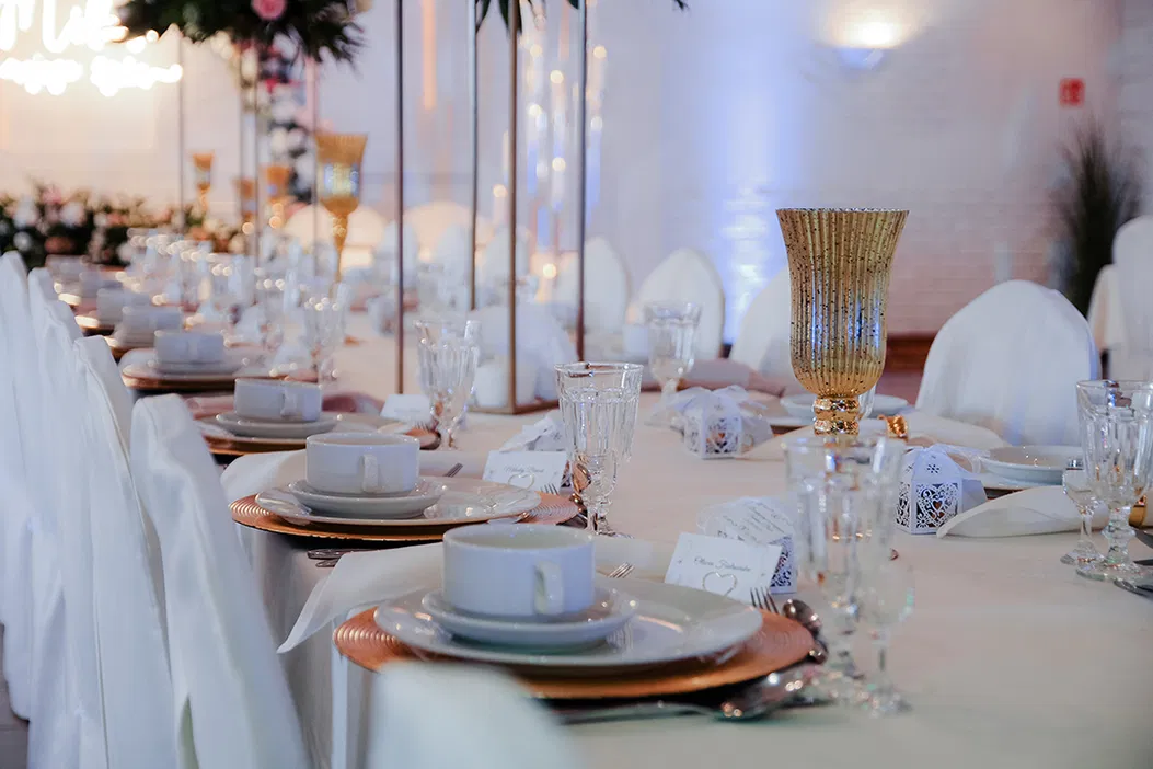 Elegancko udekorowany stół weselny z białymi pokrowcami na krzesła, złotymi akcentami i zastawą stołową, przygotowany na przyjęcie w stylu glamour.