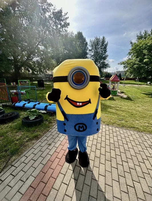 Postać w przebraniu Minionka na placu zabaw na zewnątrz, słoneczny dzień, drzewa w tle.