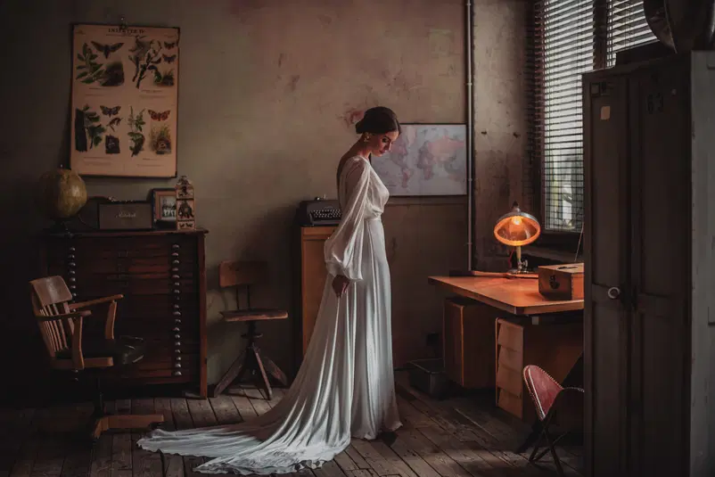  Stylowa kobieta w eleganckiej białej sukni ślubnej w przytulnym, vintageowym biurze z drewnianymi meblami i lampą na biurku. 
