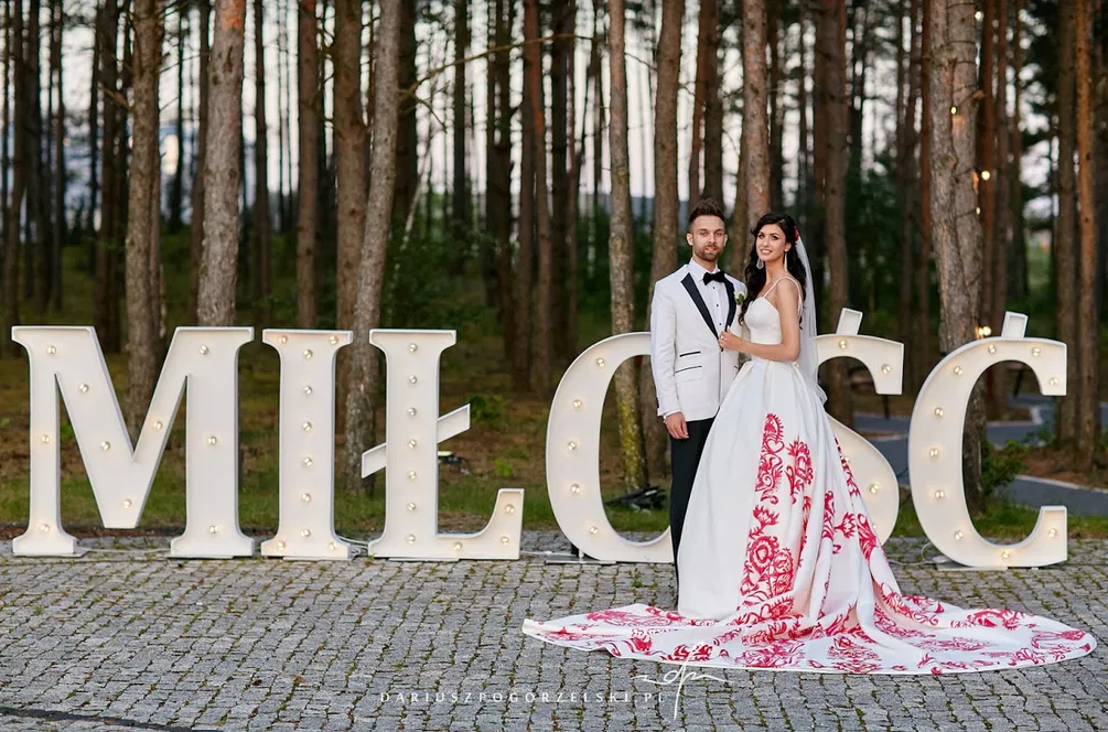 Para ślubna w lesie przed wielkimi, podświetlonymi literami tworzącymi słowo MIŁOŚĆ, panna młoda w białej sukni z czerwonymi wzorami, pan młody w białej marynarce.