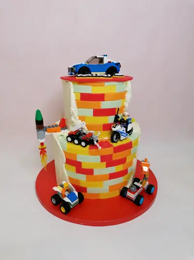  Dwupiętrowy tort dekorowany klockami LEGO i figurkami pojazdów, kolorowe wzory na bokach ciasta, na poszczególnych poziomach ustawione samochodziki i rakieta, kolorowa podstawka. 