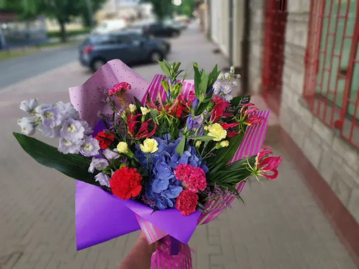  Kolorowy bukiet kwiatów owinięty w fioletowy papier, stojący na tle miejskiego chodnika. 