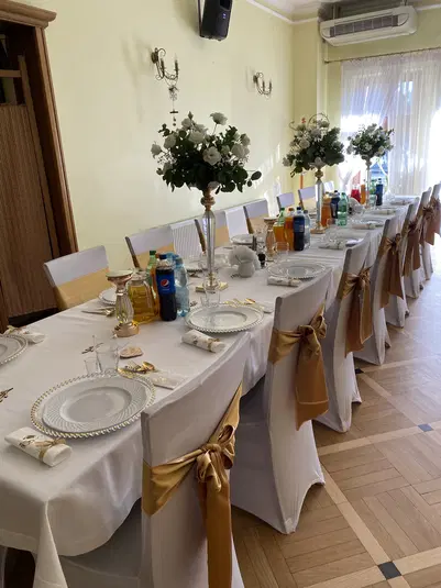  Stół weselny elegancko udekorowany, białe pokrowce na krzesła z złotymi kokardami, wysokie wazoniki z bukietami białych kwiatów, zastawa stołowa na białych obrusach, przyjęcie, sala bankietowa. 