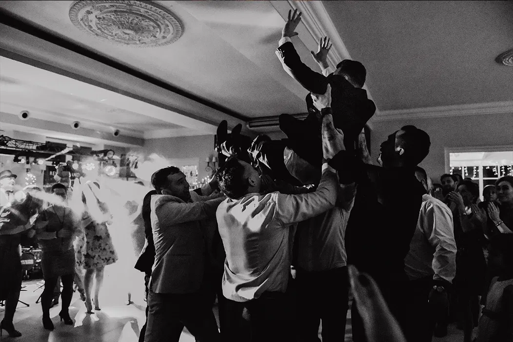 Czarno-biała fotografia przedstawiająca grupę ludzi podrzucających mężczyznę w powietrzu podczas wesela w eleganckiej sali.