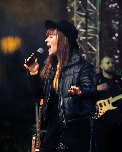 Nie wiem, kto to jest. 

Kobieta śpiewająca na scenie, ubrana w czarną kurtkę i kapelusz, trzyma mikrofon podczas koncertu na żywo. Na drugim planie widać muzyka z gitarą basową.