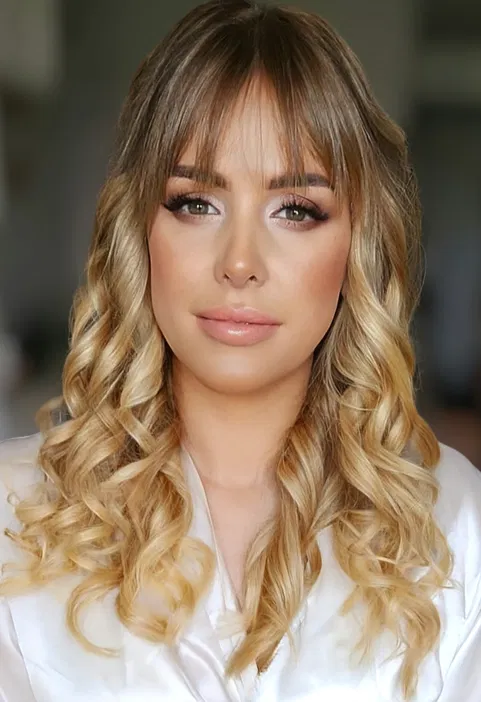 Blond kobieta z lokami i subtelnym makijażem, patrząca prosto w obiektyw, elegancko ubrana, jasne tło.
