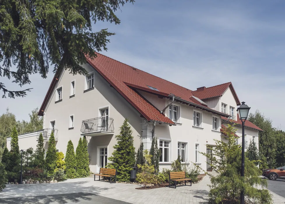 Dom z czerwonym dachem, nowoczesna architektura, zielony ogród, ławki przed budynkiem, niebieskie niebo.