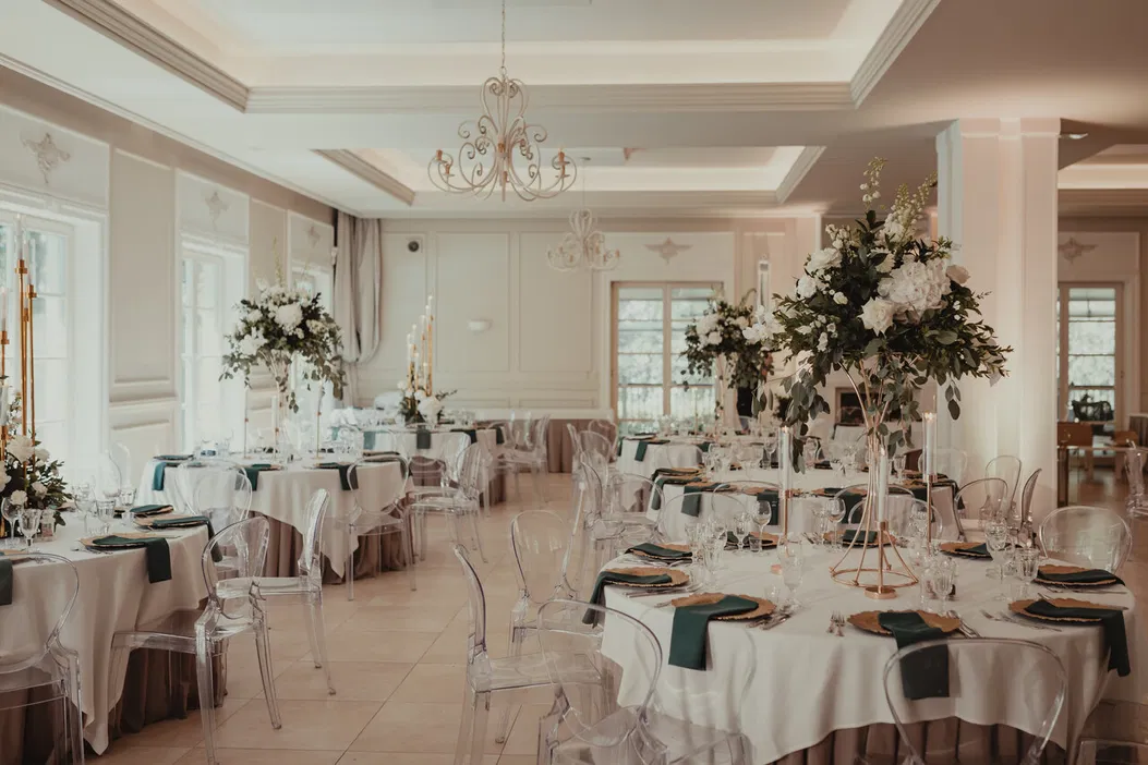 Elegancka sala weselna z dekoracjami kwiatowymi, okrągłymi stołami i przezroczystymi krzesłami, idealna na luksusową uroczystość.