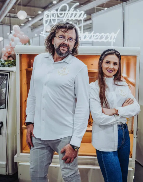 Nieznane osoby przed stylowym stoiskiem z napisem Viva Prosecco, ubrani w białe koszule, na targach branżowych.