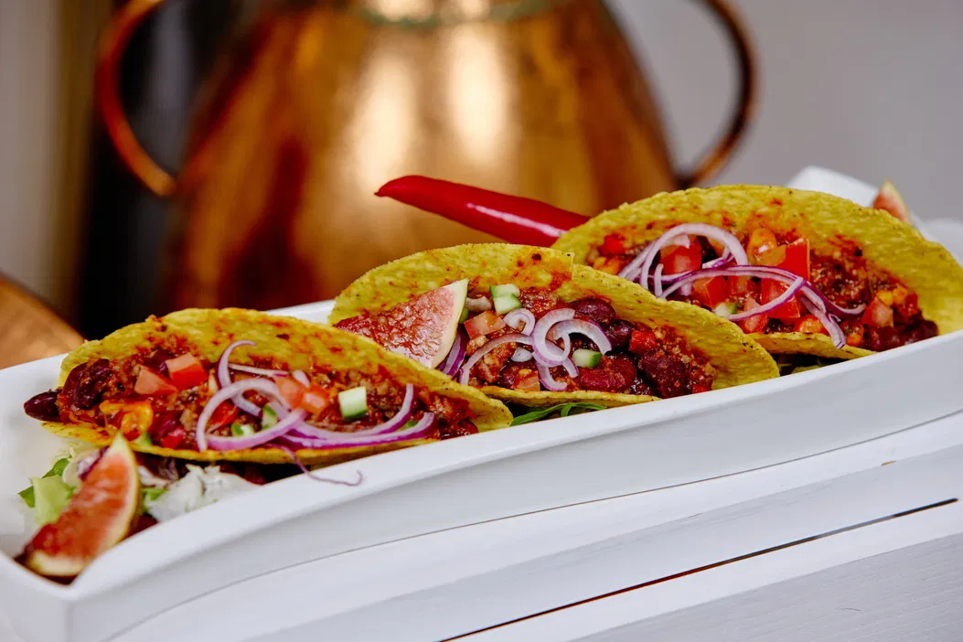 Trzy tacos z mięsem, cebulą i pomidorami na białym talerzu, udekorowane figami i ostrą papryczką, ustawione na tle miedzianego dzbanka.