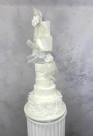 Tort weselny z białymi dekoracjami w stylu glamour, ozdobiony falbankami i perłami na tle jasnej ściany. Wysoki, elegancki tort piętrowy idealny na uroczystość weselną.