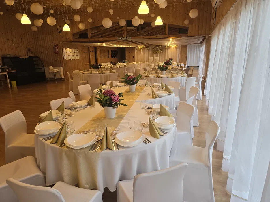 Elegancka sala bankietowa z okrągłymi stołami udekorowanymi białymi obrusami i złotymi serwetkami, przygotowana na przyjęcie weselne.