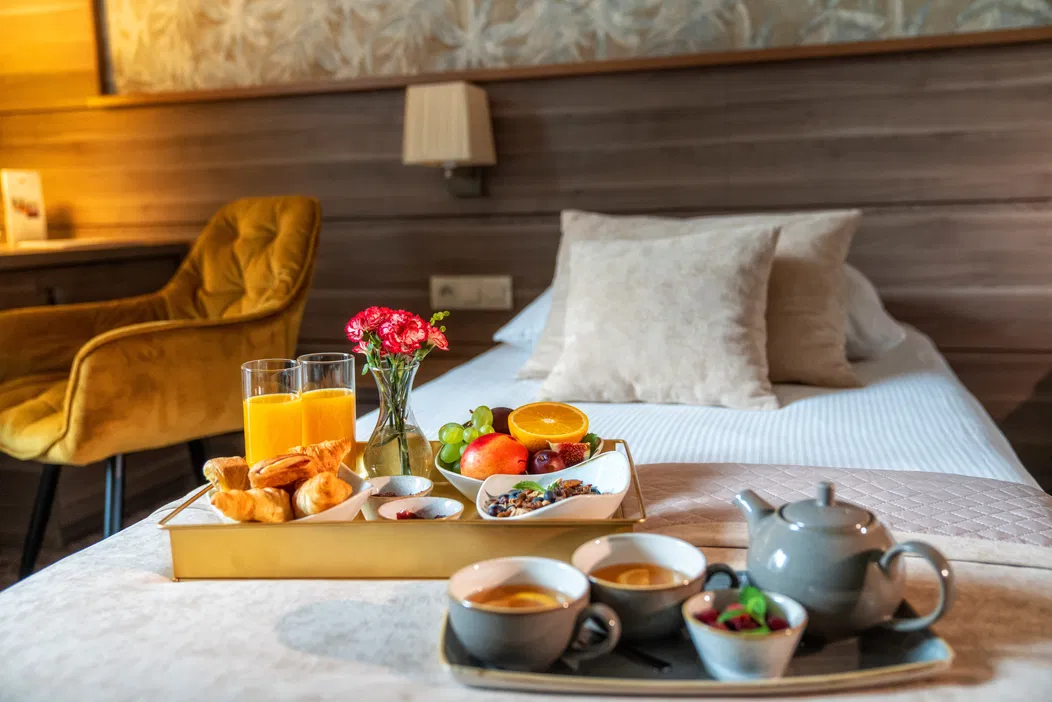 Luksusowy hotelowy pokój z podanym śniadaniem na tacy, w tym świeżo wyciskany sok pomarańczowy, owoce, rogaliki i herbata na elegancko pościelonym łóżku. Idealne miejsce na relaks i komfortowy pobyt.