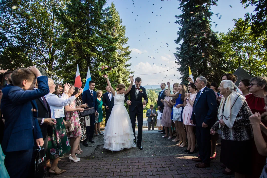 Para młoda opuszczająca ceremonię ślubną, otoczona przez gości weselnych i przyjaciół, z radością i uśmiechami w świetle słonecznym, flagi narodowe w tle, uroczystość na świeżym powietrzu, radosny moment.