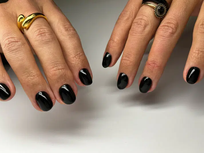 Czarne paznokcie hybrydowe na dłoniach z pierścionkami, elegancki manicure na jasnym tle. 