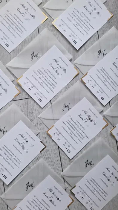 Eleganckie zaproszenia ślubne z inicjałami AK i złotymi zdobieniami, na białym tle drewnianej podłogi.