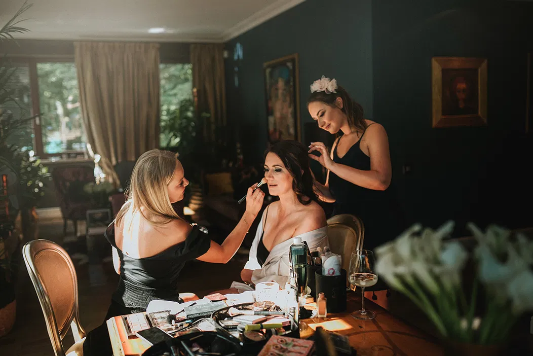 Makeup artist applying makeup to a woman while another woman styles her hair in a warmly lit room with elegant decor, flowers and a wine glass nearby.
