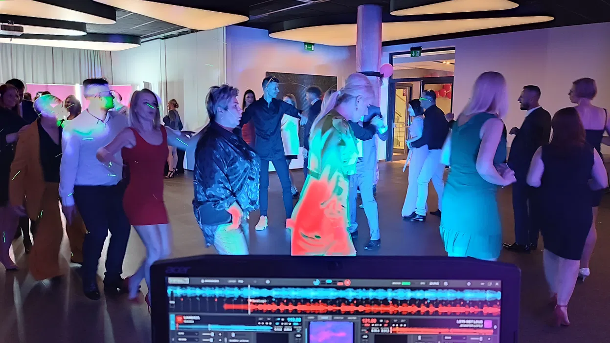 Ludzie tańczący na przyjęciu, kolorowe światła LED, nowoczesne wnętrze, widok z DJ-skiej konsolety, zabawa taneczna na imprezie, elegancko ubrani uczestnicy.