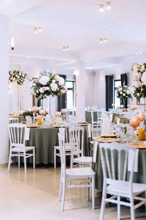 Elegancka sala weselna z dekoracją kwiatową, okrągłymi stołami i białymi krzesłami, przygotowana na przyjęcie.