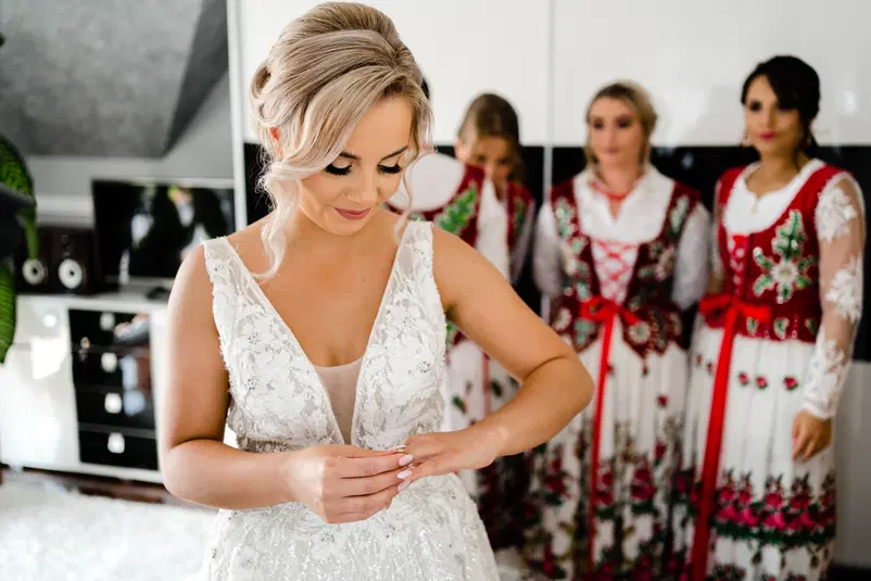  Panna młoda w białej sukni ślubnej, przygotowująca się do ceremonii, otoczona druhnami w tradycyjnych strojach ludowych. 