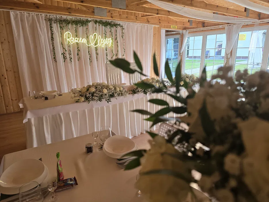 Dekoracja stołu weselnego z napisem Razem lepiej, elegancka sala z białymi zasłonami i kwiatami, przygotowana na uroczystość ślubną.