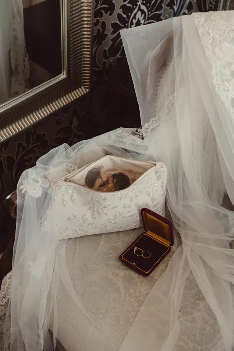 Biała koronkowa torebka ślubna z fotografią pary, obok których leży etui z obrączkami, na tle welonu i eleganckiego krzesła.