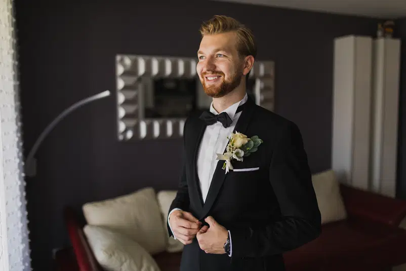  Mężczyzna w eleganckim garniturze z muchą, z przypiętą butonierką, uśmiechający się w jasnym pomieszczeniu przed lustrem. 