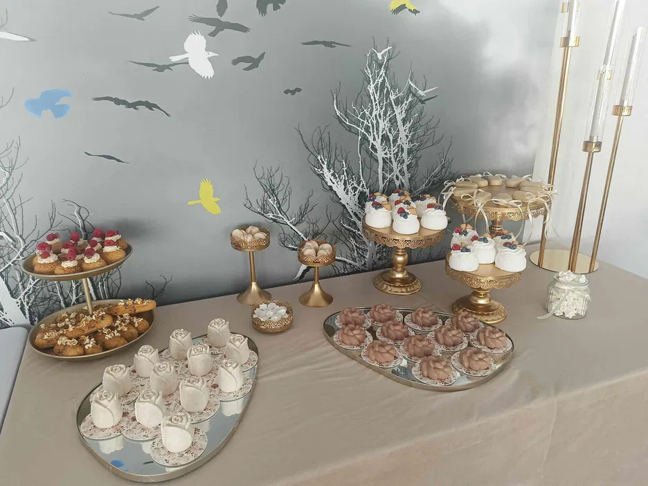 Stół deserowy z ciastkami i bezami ozdobionymi owocami, ustawionymi na eleganckich złotych paterach, przygotowany na uroczystość.