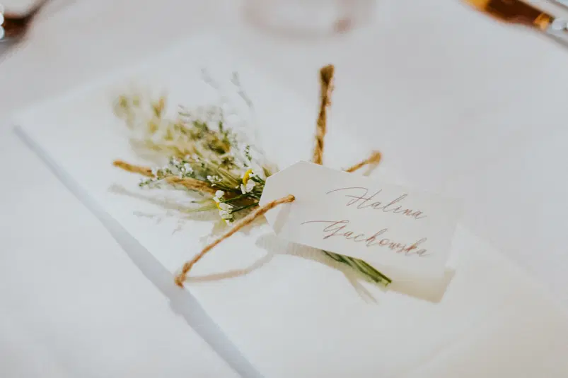  Rustykalna dekoracja ślubna z gałązkami i personalizowaną winietką на białym obrusie. 