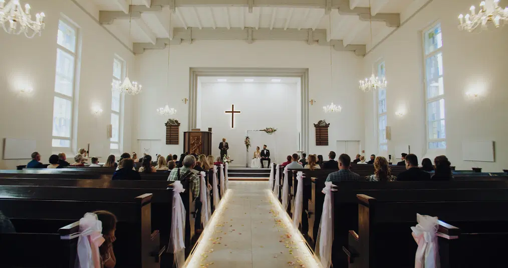  Ceremonia ślubna w kościele, para młoda na ołtarzu, goście siedzący w ławkach, dekoracje z tkaniny i kwiatów. 
