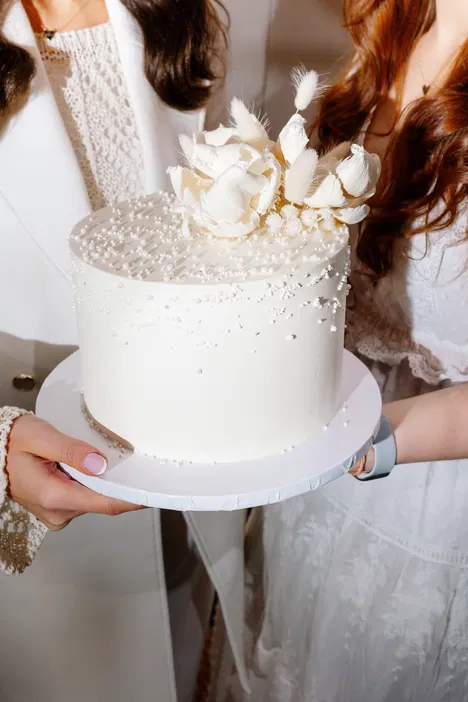 Biały tort ślubny ozdobiony kwiatami, trzymany przez dwie osoby w eleganckich strojach.