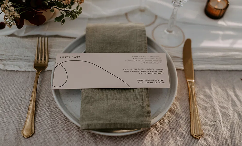 Elegancka zastawa stołowa z menu na talerzu, zielona serwetka, złote sztućce, kwiaty w wazonie, nastrojowe oświetlenie.