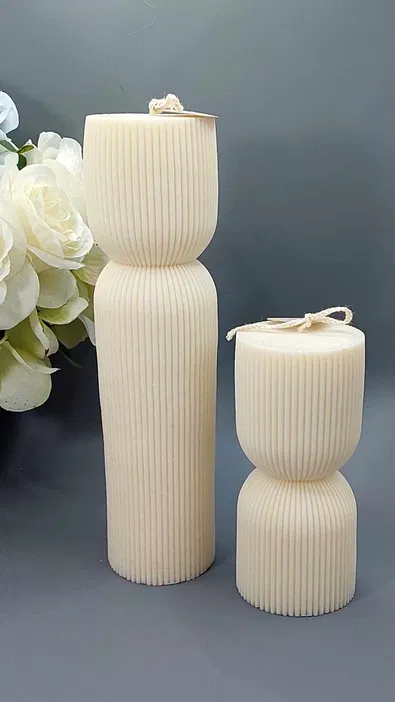 Dwie białe, żebrowane świece sojowe o różnych rozmiarach, stojące na tle szarych kwiatów, dekoracyjne świece handmade.