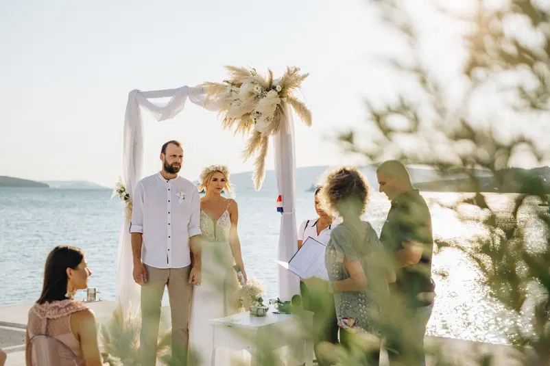  Ślub na plaży z widokiem na morze, ceremonia pod baldachimem z białych tkanin i ozdobą z traw, para młoda i goście w letnich strojach, słońce zachodzące nad wodą. 