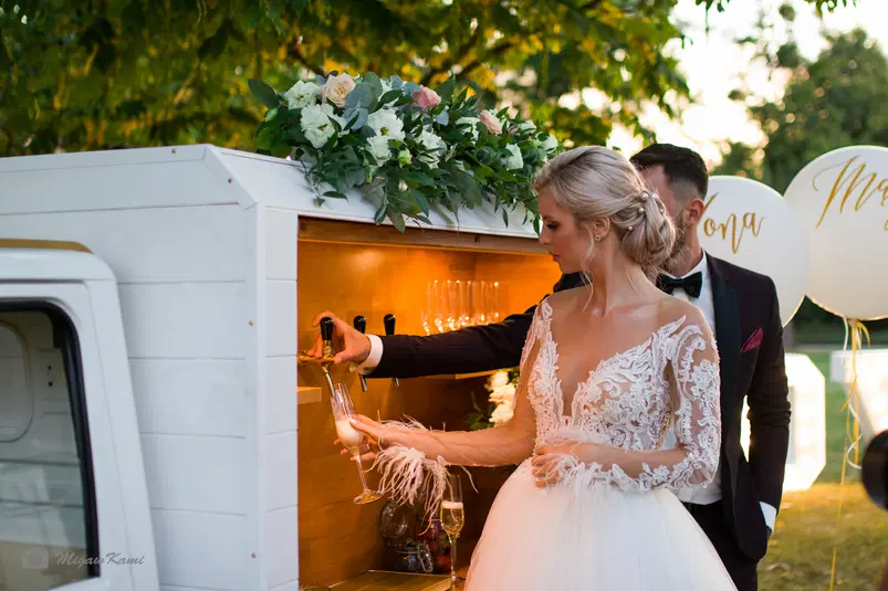  Para młoda przy barze plenerowym, elegancka panna młoda w koronkowej sukni ślubnej, mężczyzna w garniturze nalewający szampana na przyjęciu weselnym na świeżym powietrzu. 
